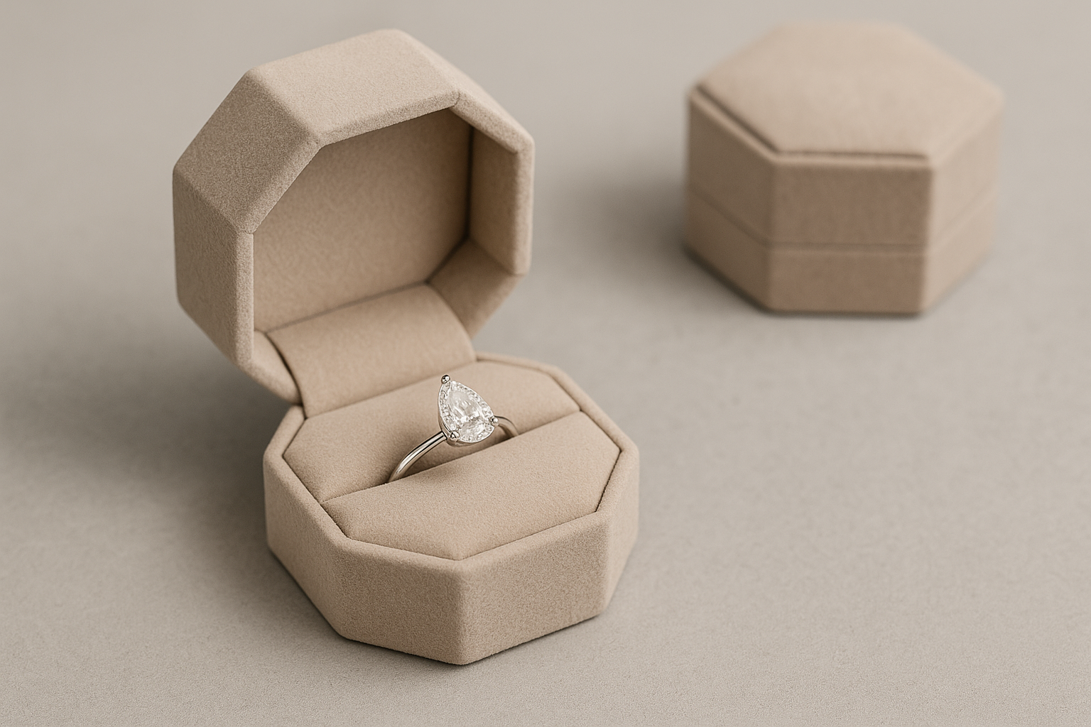 Išskirtinis sužadėtuvių žiedo įpakavimas – atvira šešiakampė smėlio spalvos dėžutė su lašo formos deimantiniu žiedu, fone uždaryta tokia pati dėžutė