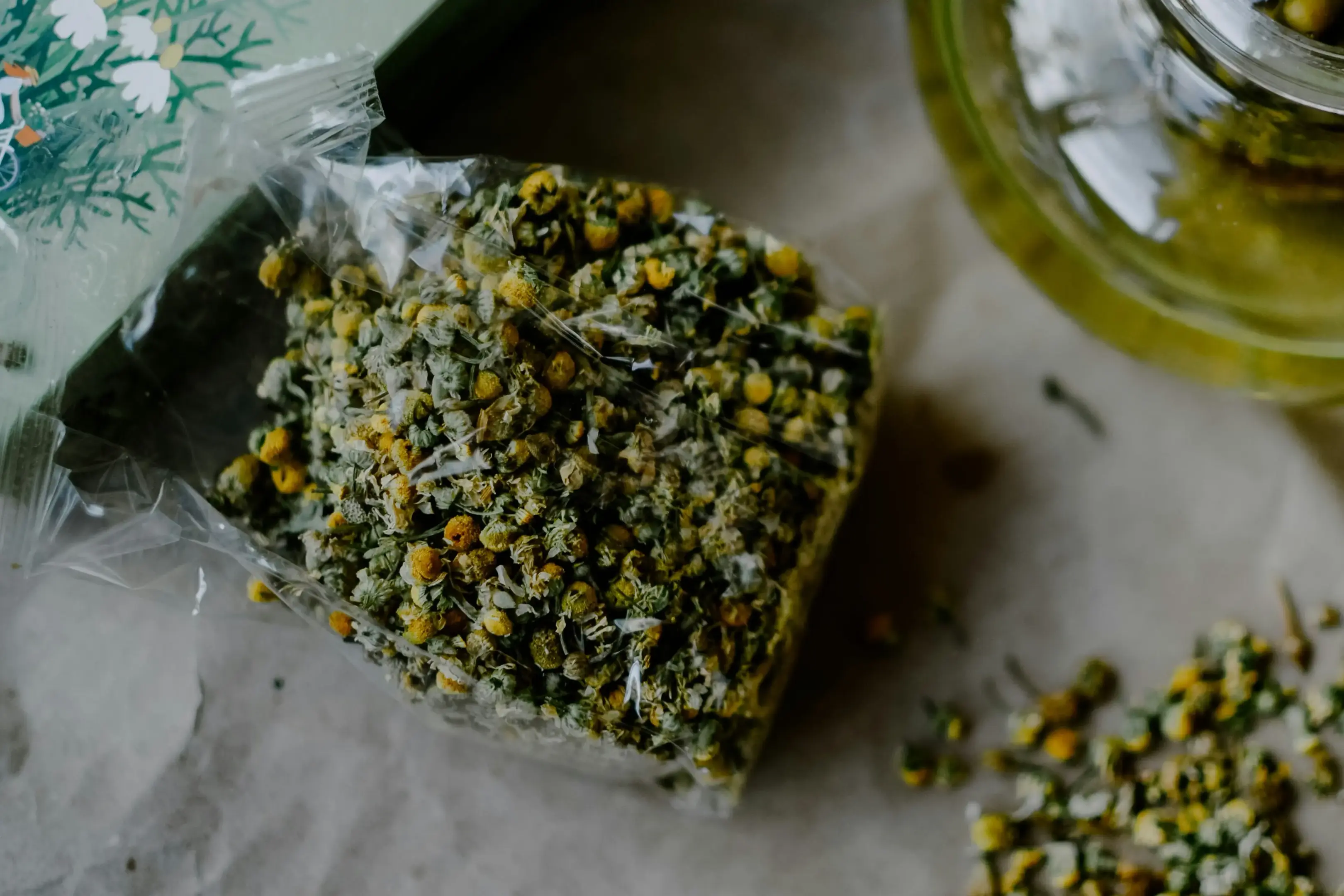 A clear plastic bag filled with dried chamomile flowers sits on a textured, neutral-toned surface next to a glass teapot. A small pile of loose, dried chamomile buds is scattered on the lower right side of the frame.
