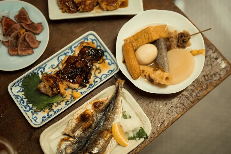 A rustic wooden table displaying a hearty Japanese meal, including a whole grilled fish, a plate of savory glazed chicken, sliced seared meat, and a comforting bowl of oden.