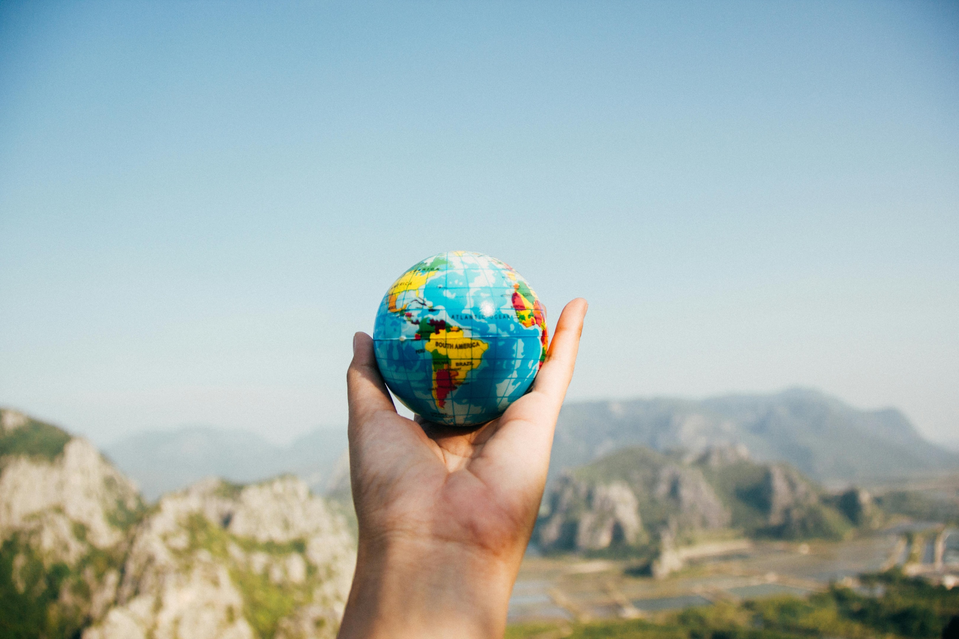 Hand holding a small globe with a mountainous landscape and clear blue sky in the background, symbolizing travel and global exploration.