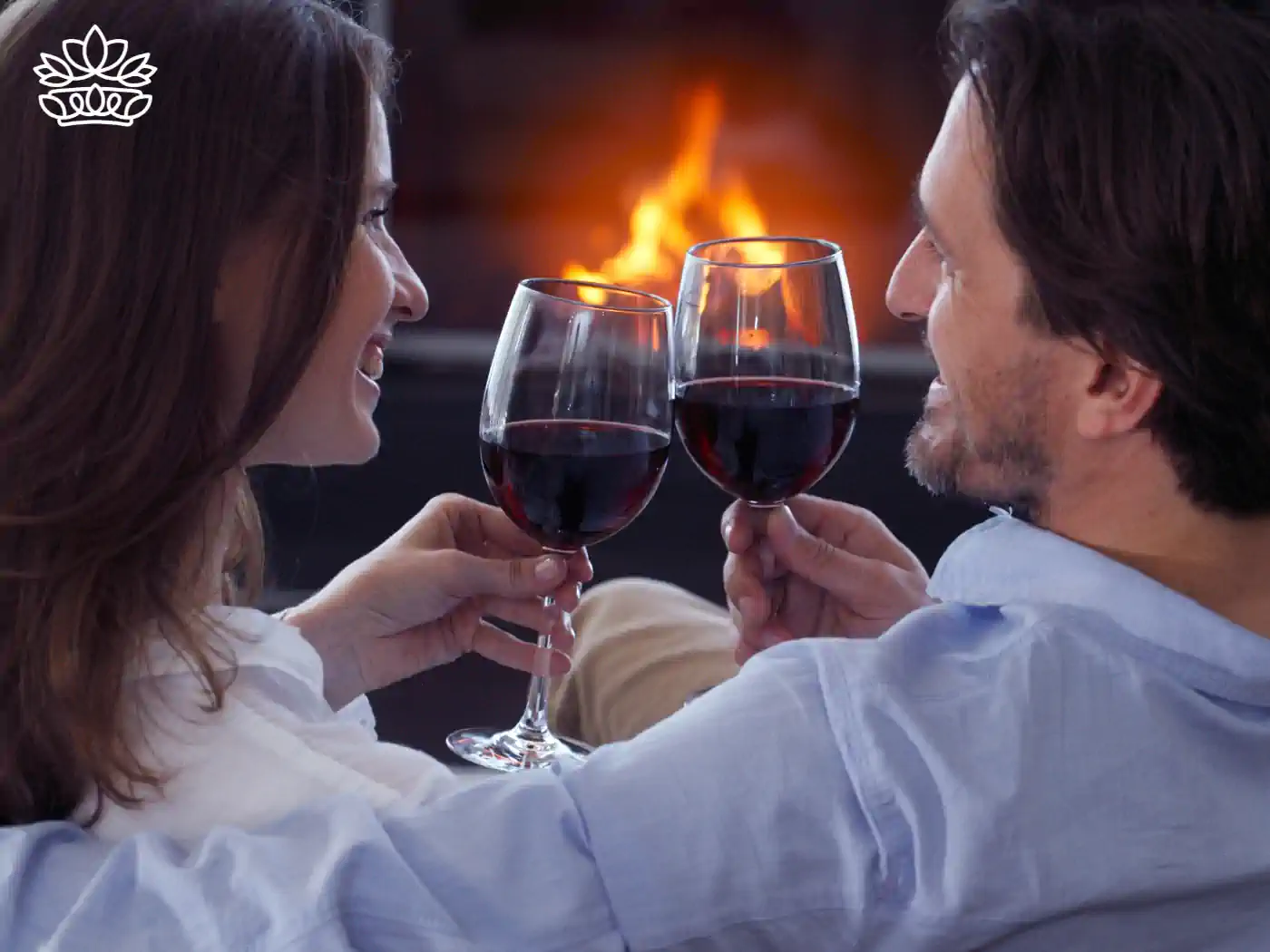 Couple clinking red wine glasses in front of a fireplace - Fabulous Flowers and Gifts: Gift Boxes with Champagne and Wine.