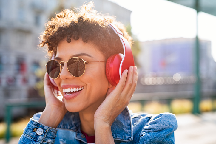 Happy young woman with short dark hair and sunglasses listening to music through red headphones.