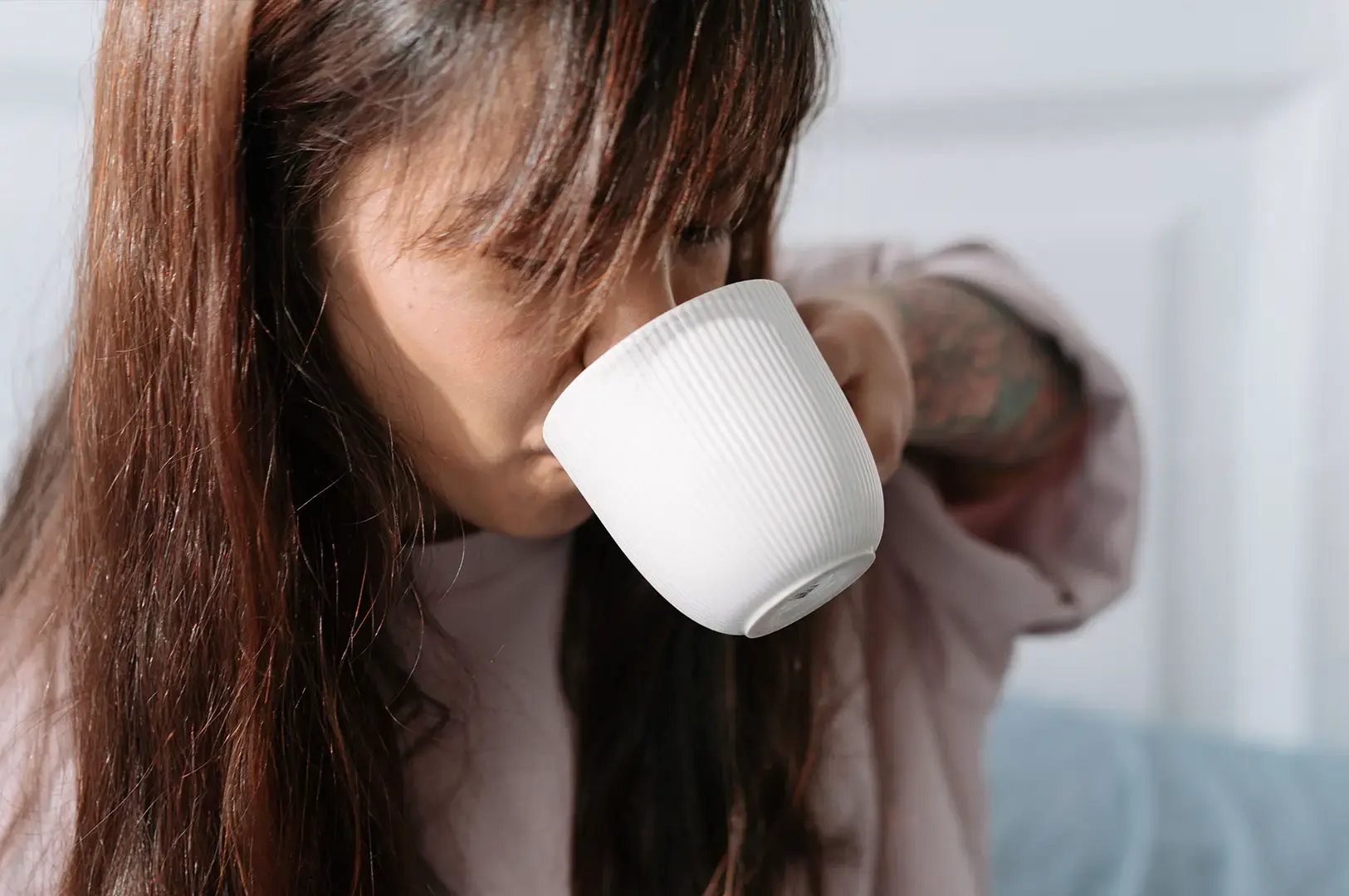A close-up, shallow-depth-of-field shot shows a person with long brown hair taking a sip from a white ceramic mug. The mug is decorated with fine, vertical textured ridges along its exterior. The focus is sharp on the mug and the person's hand holding it, while their facial features and a soft white paneled wall in the background remain gently out of focus.