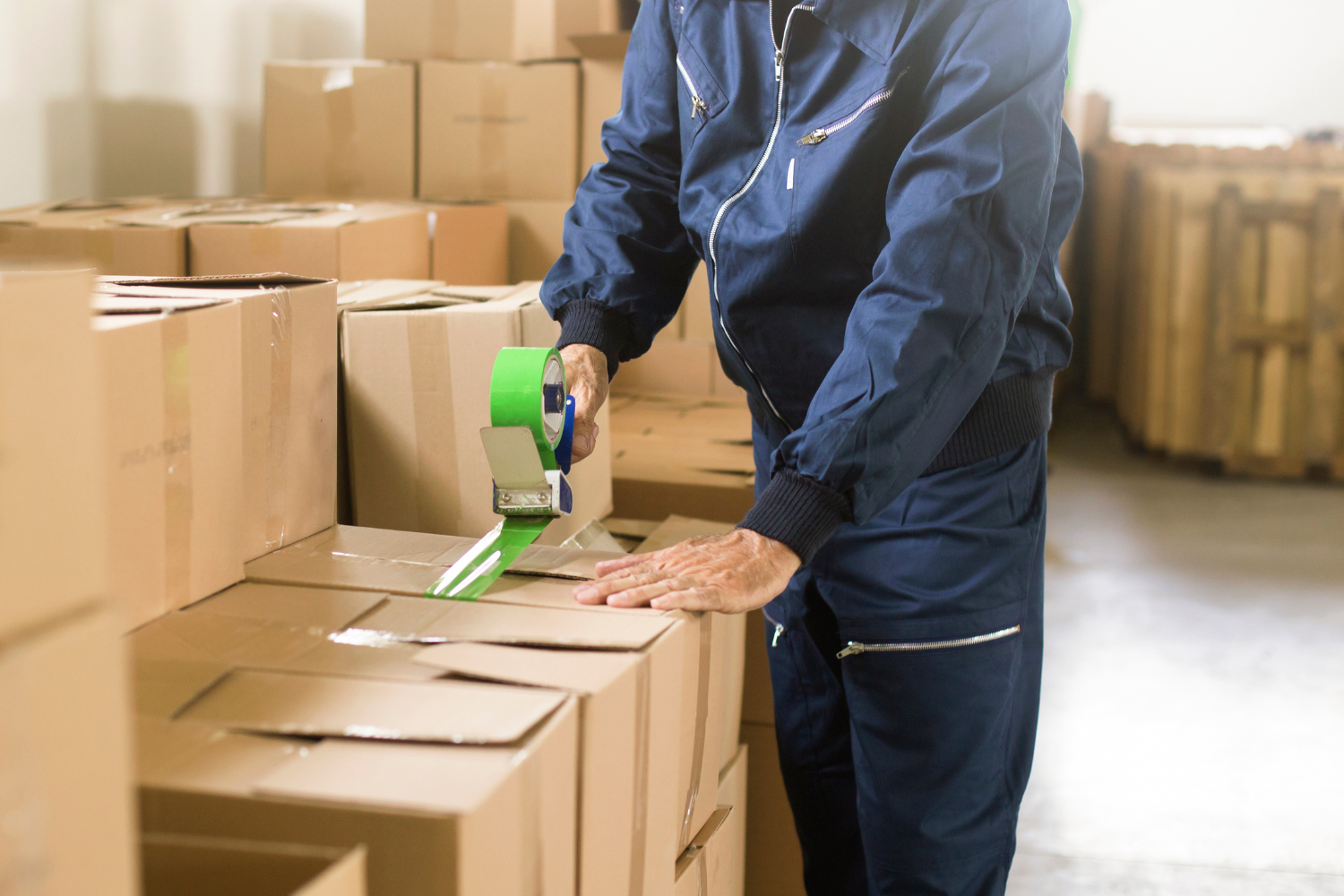 A person in a blue jumpsuit tapes shut cardboard boxes in a warehouse.