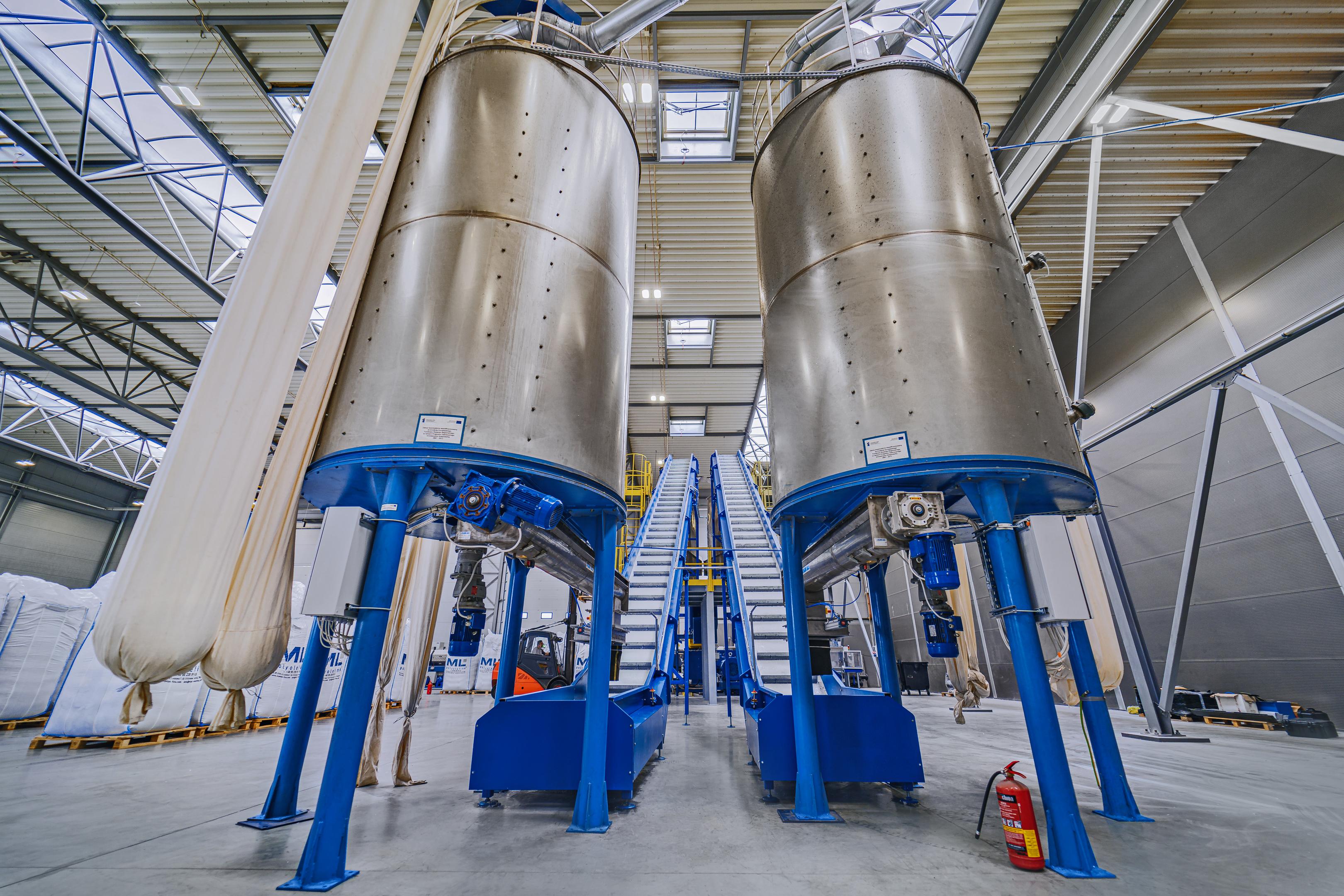 Na obrazie przedstawiona jest linia produkcyjna do recyklingu tworzyw sztucznych, wyposażona w taśmociąg oraz ekstruder. Proces ten przyczynia się do ochrony środowiska, umożliwiając przetwarzanie odpadów z tworzyw sztucznych, takich jak butelki i opakowania, w granulat, który znajduje zastosowanie w produkcji elementów samochodowych oraz rur.