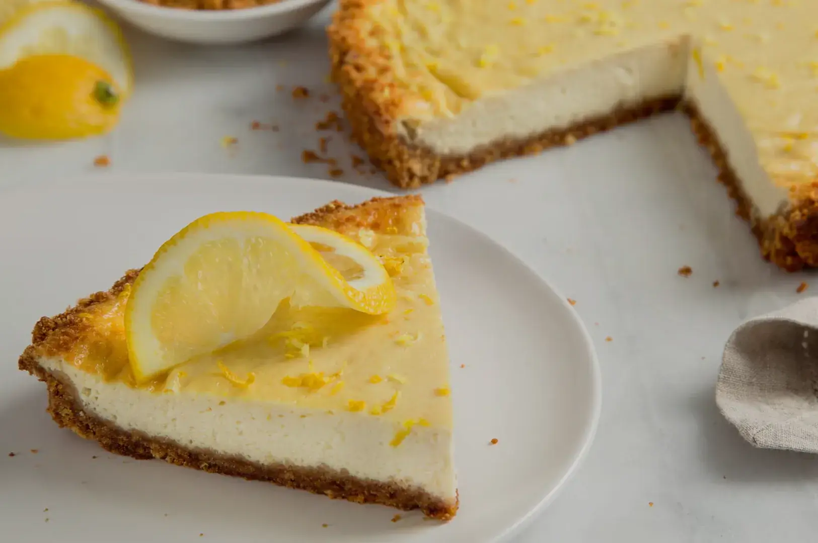 A slice of lemon cheesecake on a white plate, garnished with a lemon slice, with the pie partially cut and visible in the background.