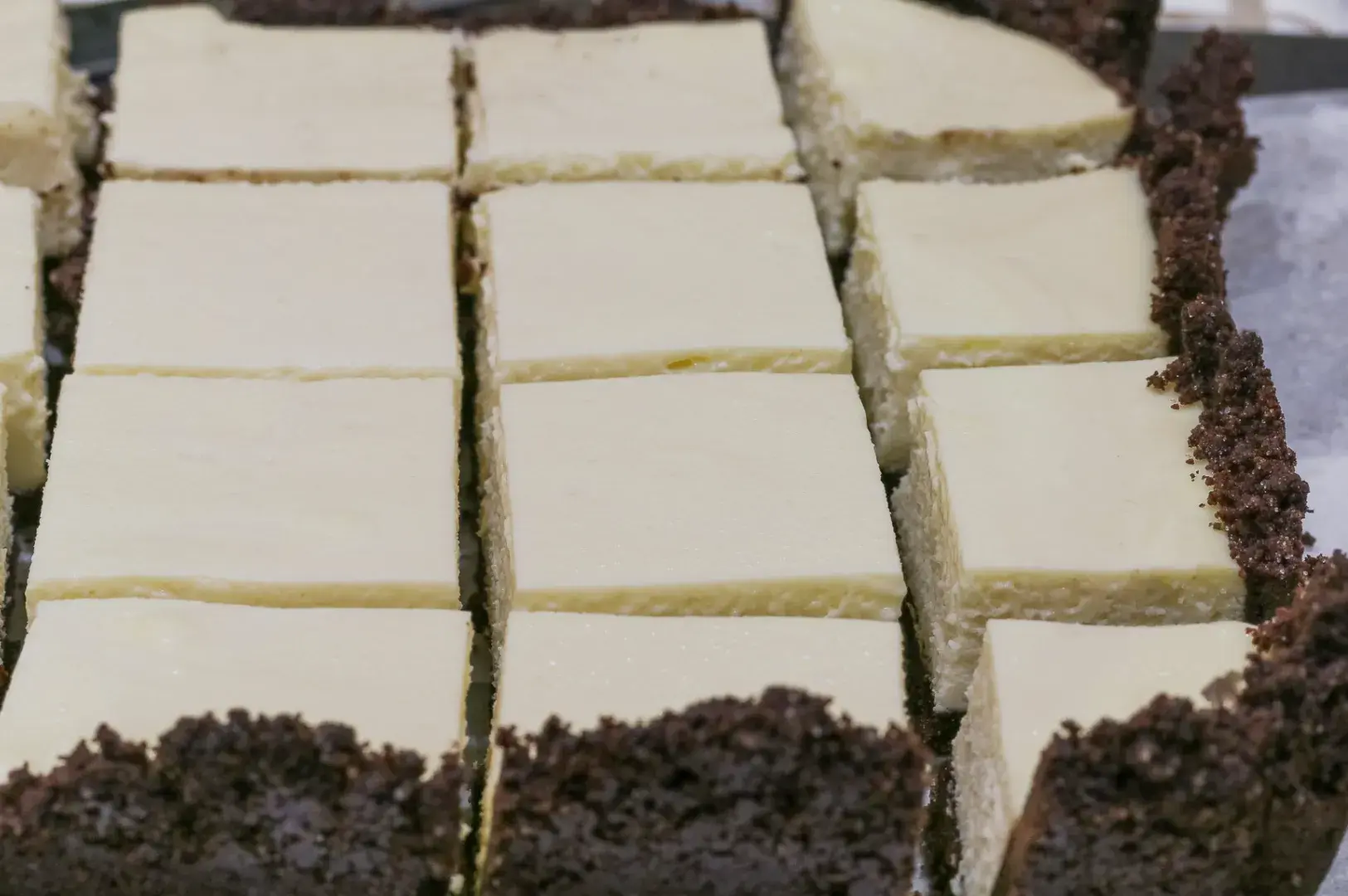 Rows of creamy white dessert bars with dark chocolate-cookie crust, neatly cut in a tray; the close-up looks rich and inviting.