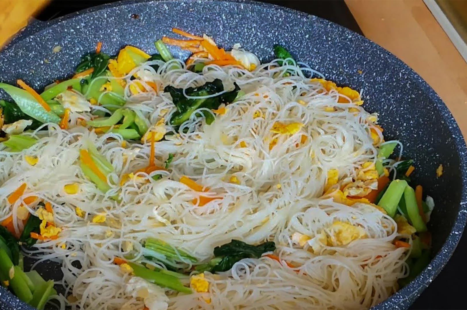 A close-up shot of rice vermicelli being cooked in a large non-stick pan with scrambled eggs and green vegetables.