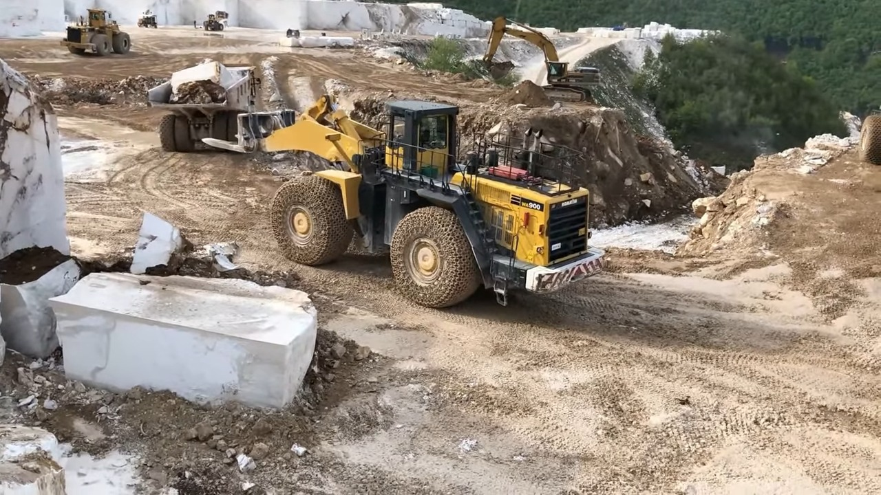 A yellow and black wheel loader carrying a large block of marble in a quarry with other construction equipment and piles of marble blocks in the background
