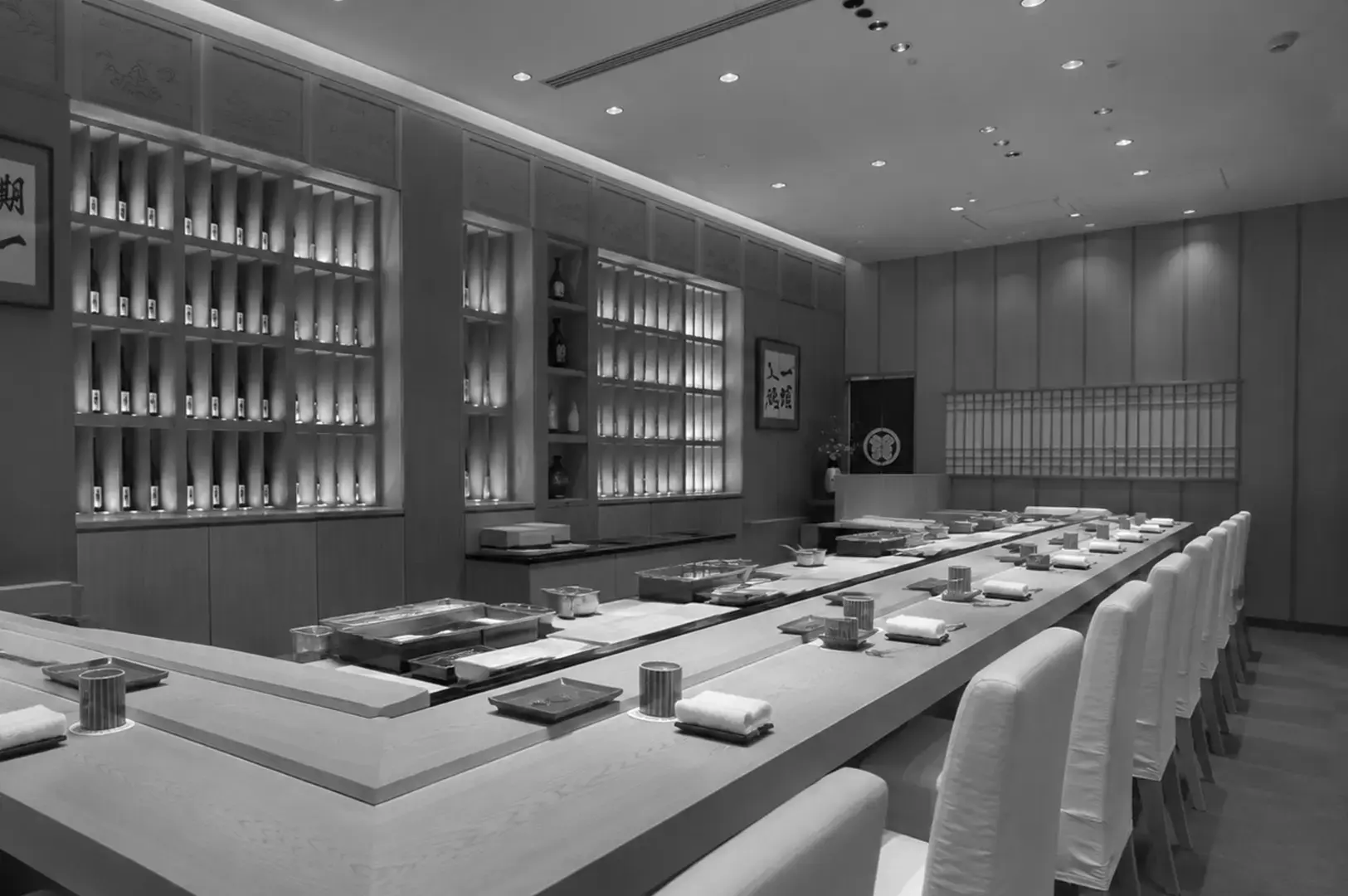 Spacious Japanese restaurant interior with a long sushi counter, white chairs, illuminated bottle displays, and dark stone walls.