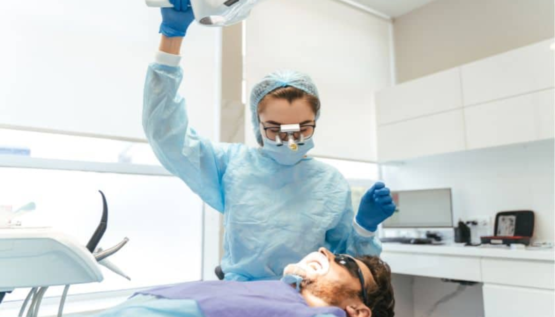 Helfo: Slik får du støtte til tannbehandling 9 Tannlege i fullt verneutstyr justerer operasjonslampe over pasient som ligger i stolen med beskyttelsesbriller.