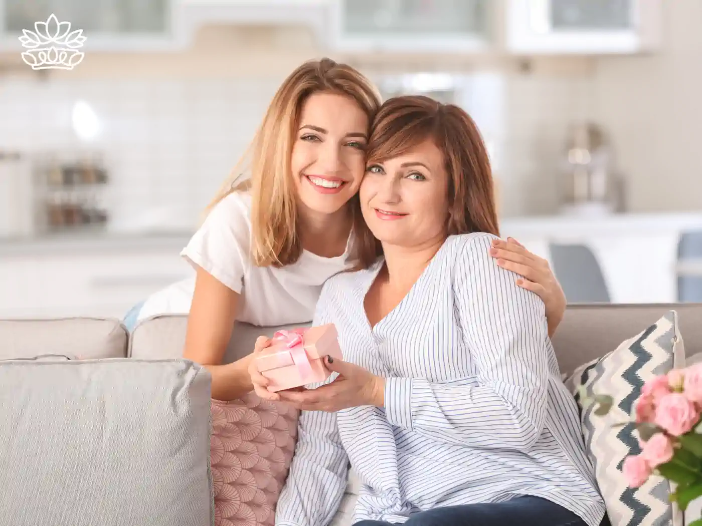 A mother and daughter sitting on a sofa, holding a pink gift box together. Fabulous Flowers and Gifts - Luxury Cape Town Gift Hampers.