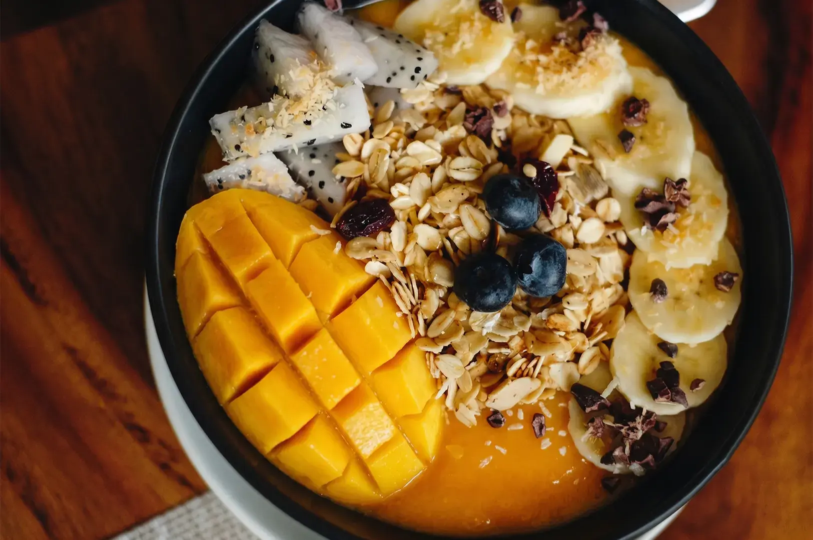 A vibrant smoothie bowl topped with fresh mango, banana slices, dragon fruit, blueberries, and crunchy granola.