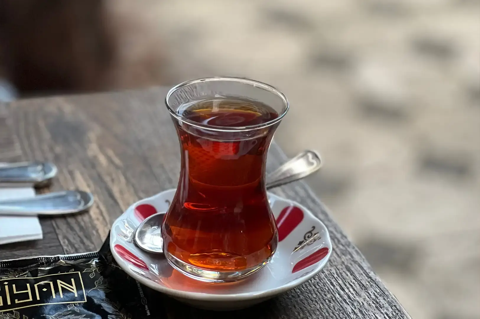 A tulip-shaped glass of dark red Turkish tea sits on a traditional white ceramic saucer adorned with red accent marks, accompanied by a small metal spoon resting on the saucer beside a packaged tea item on a rustic wooden table.