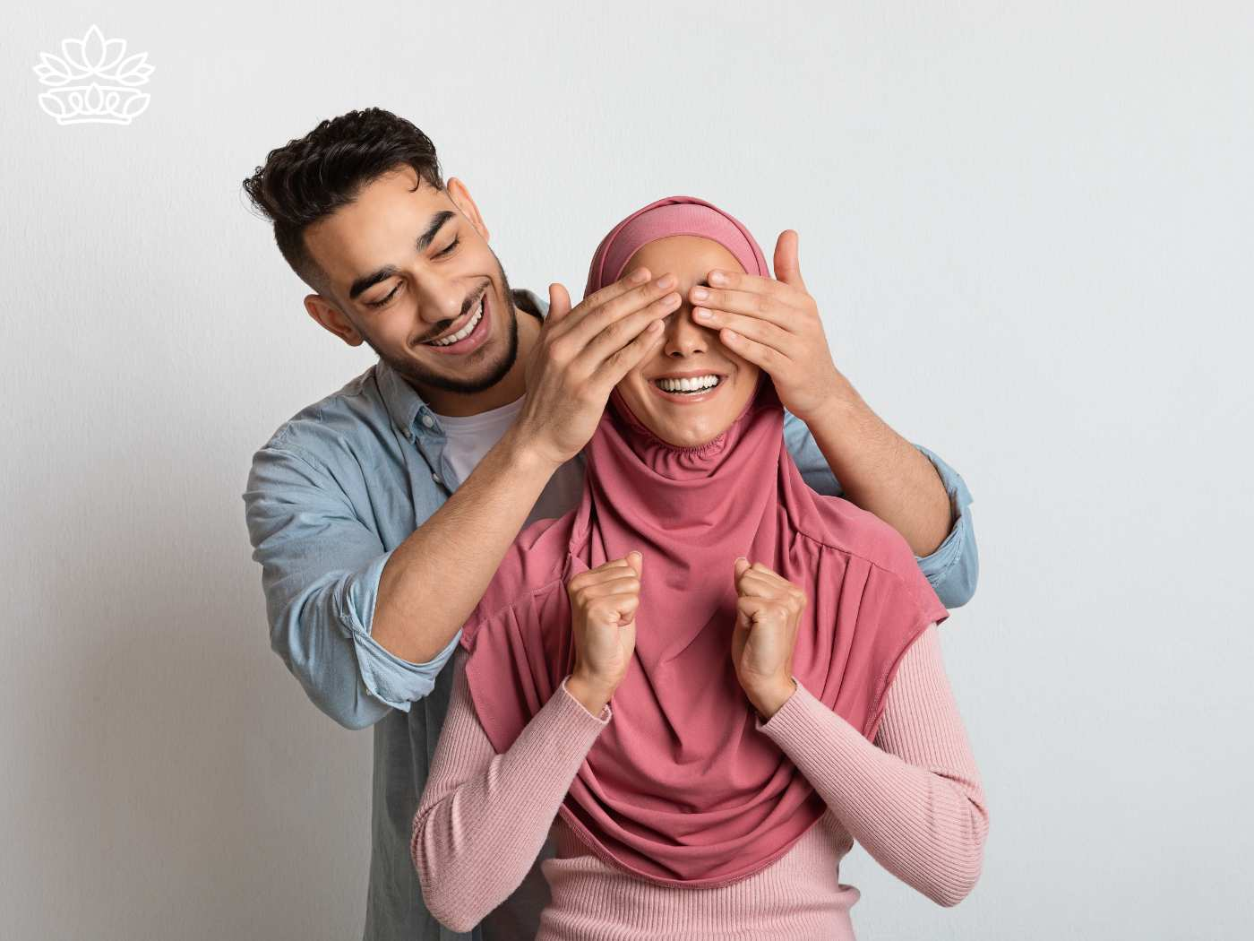 A cheerful man covering the eyes of a smiling woman wearing a pink hijab from behind in a playful gesture of surprise against a light background. Both exhibit expressions of joy and excitement, perfect for special occasions with Fabulous Flowers and Gifts. Gift Boxes for wife. Delivered with Heart.
