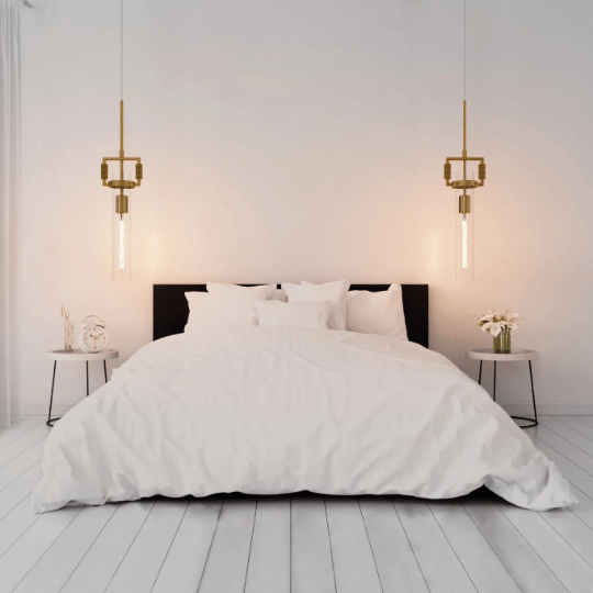 Minimalist bedroom with a large bed centered between two round side tables, each illuminated by a hanging brass pendant light with vertical glass tubes, casting a warm glow on the white walls.