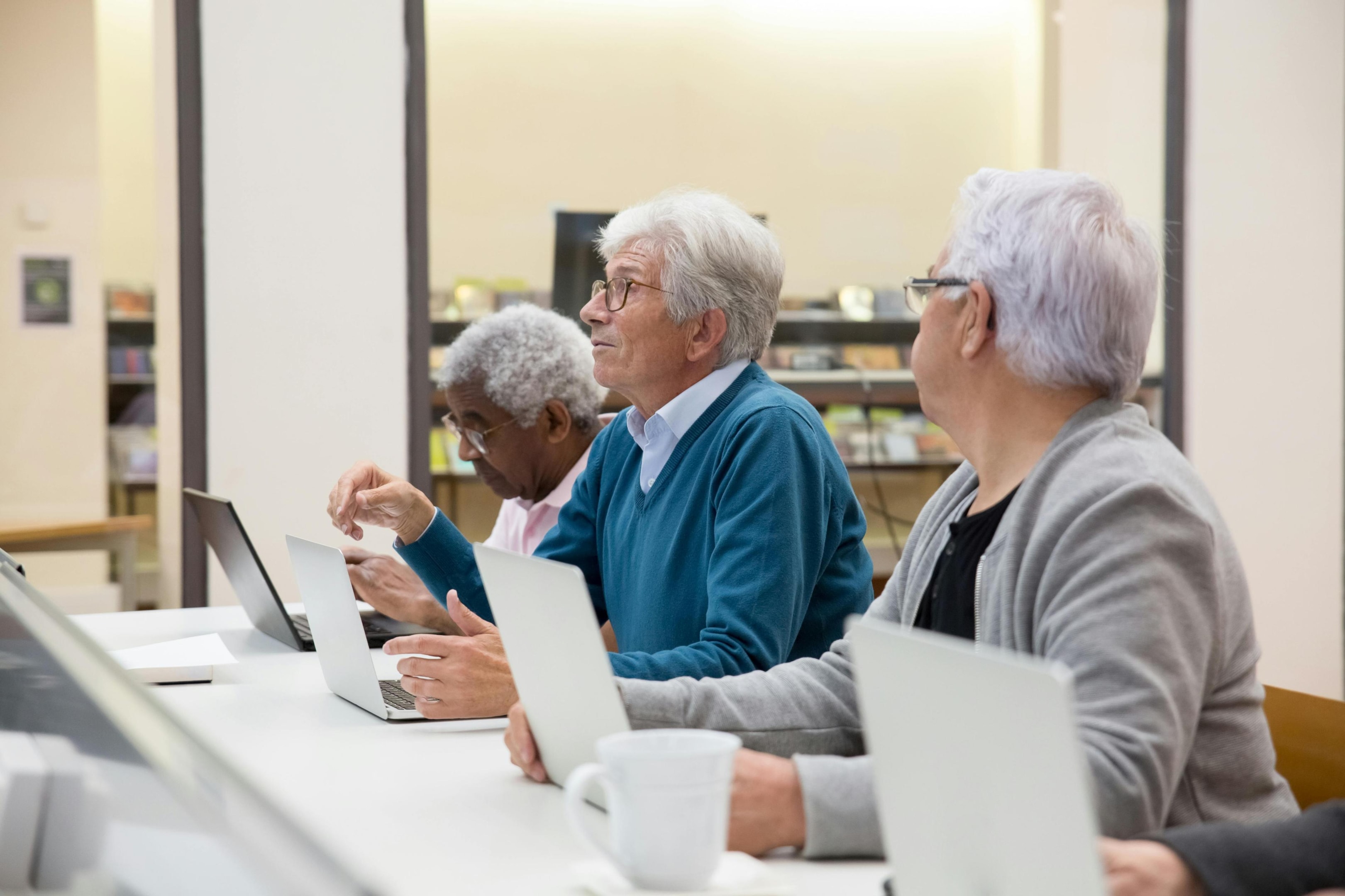This image depicts a group of senior men sitting in a row at a white table, utilizing laptops in a bright, modern space that suggests a library or classroom. While most are focused on their screens or typing, one man looks up attentively, highlighting an atmosphere of active learning and digital engagement.