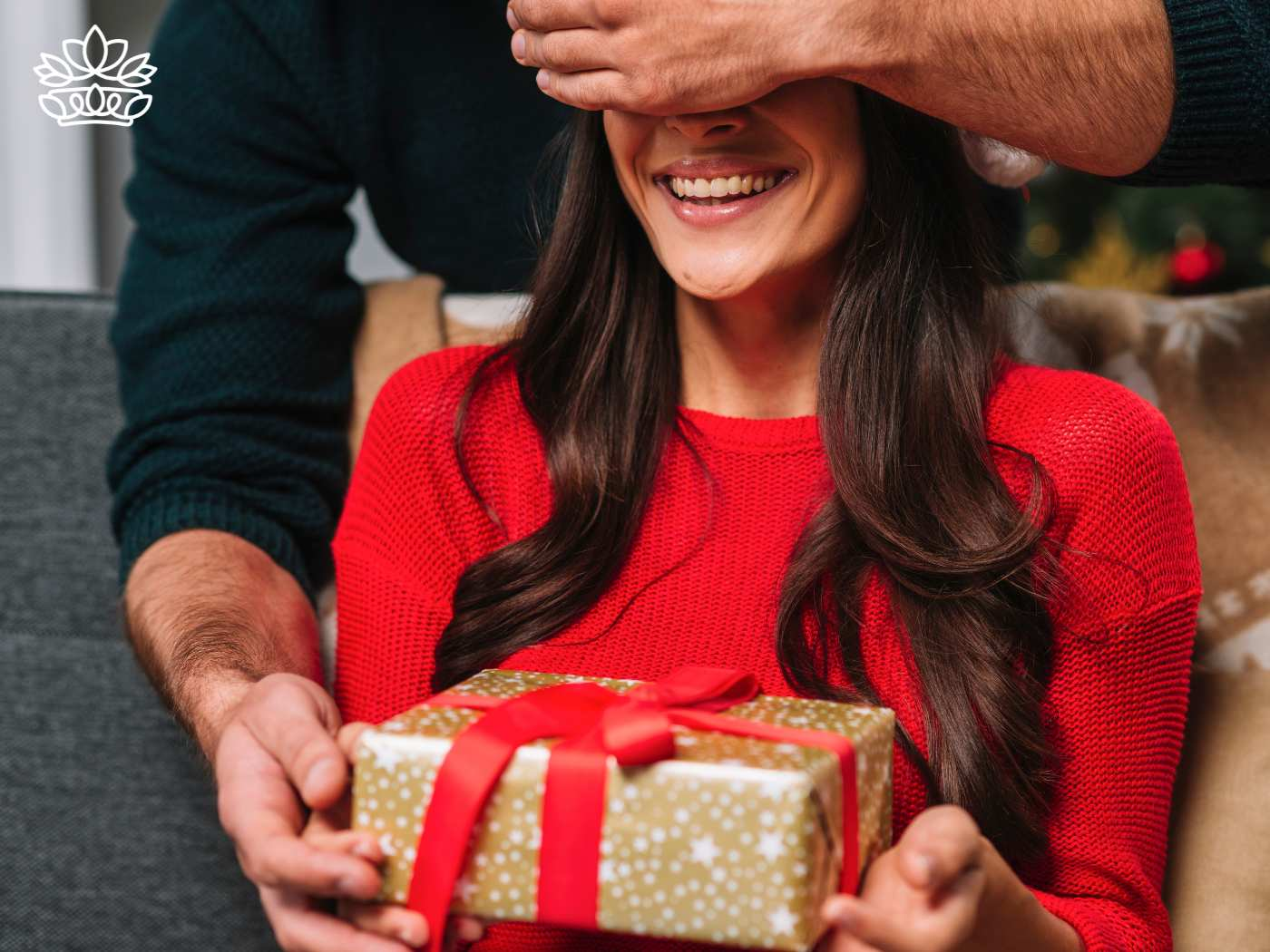 A woman in a red sweater being joyfully surprised with a gift box wrapped in festive paper, representing the Gift Boxes for Her Collection - Nationwide delivery with gift ideas - Fabulous Flowers and Gifts