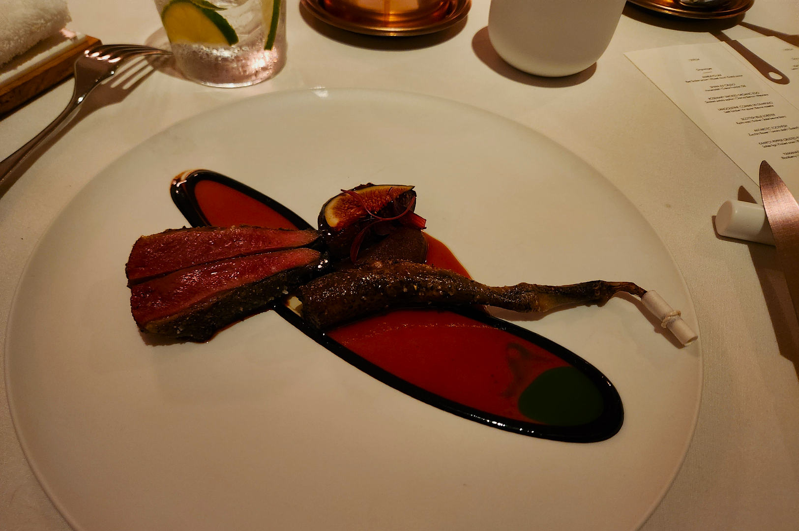 Elegant dish on a white plate featuring sliced meat, fig, and garnishes, artistically drizzled with colorful sauces, creating a gourmet presentation.
