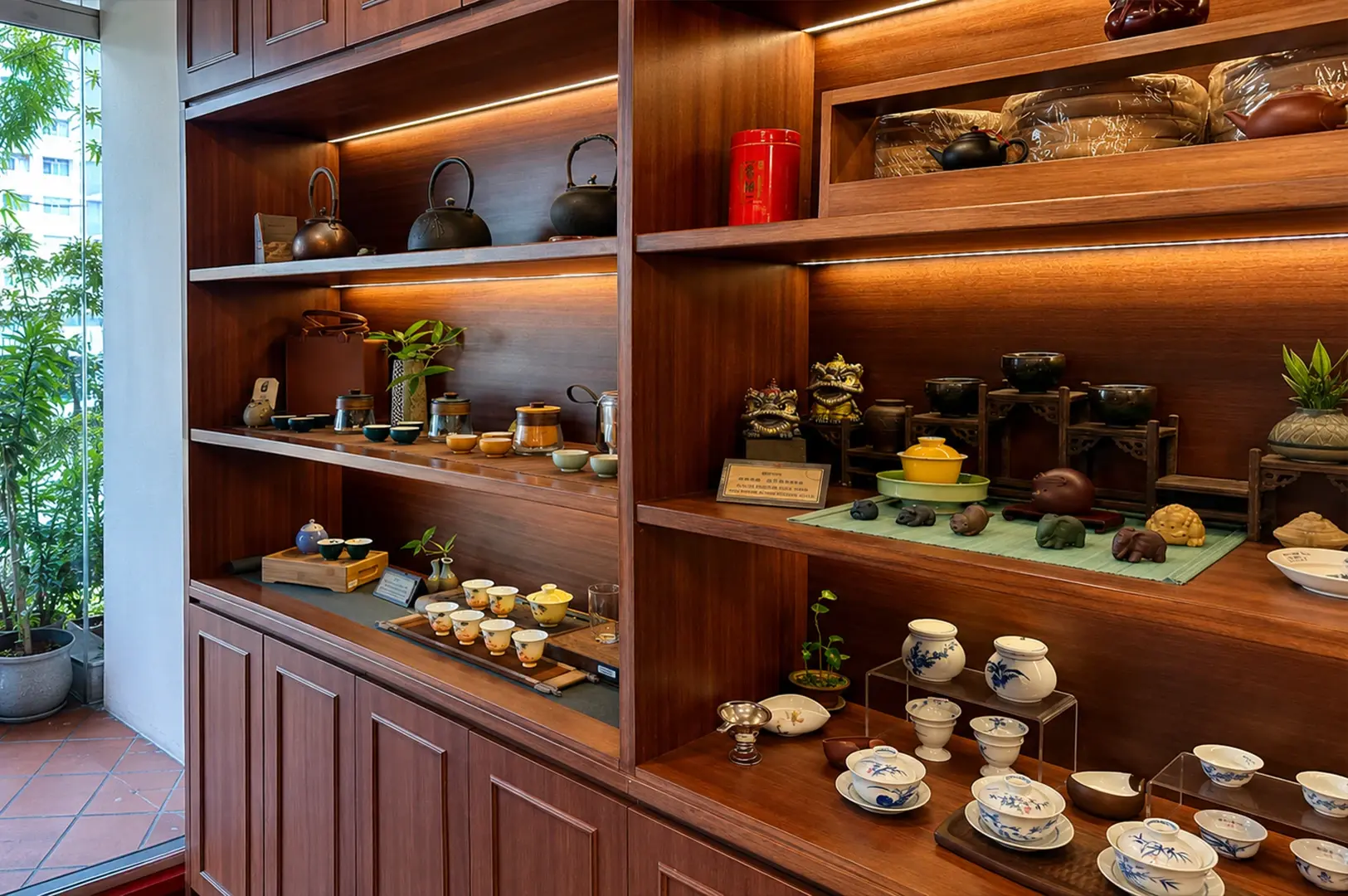 A tall, elegant wooden shelving unit is filled with an extensive collection of artisanal tea ware, including cast-iron kettles, small ceramic cups, teapots, and decorative figurines. The shelves are illuminated by soft, integrated lighting that highlights the craftsmanship of the items, which are arranged in organized, aesthetic displays alongside small potted plants. To the left, a large glass door opens to a view of green foliage, adding a natural, bright touch to the sophisticated and warm ambiance of the room.