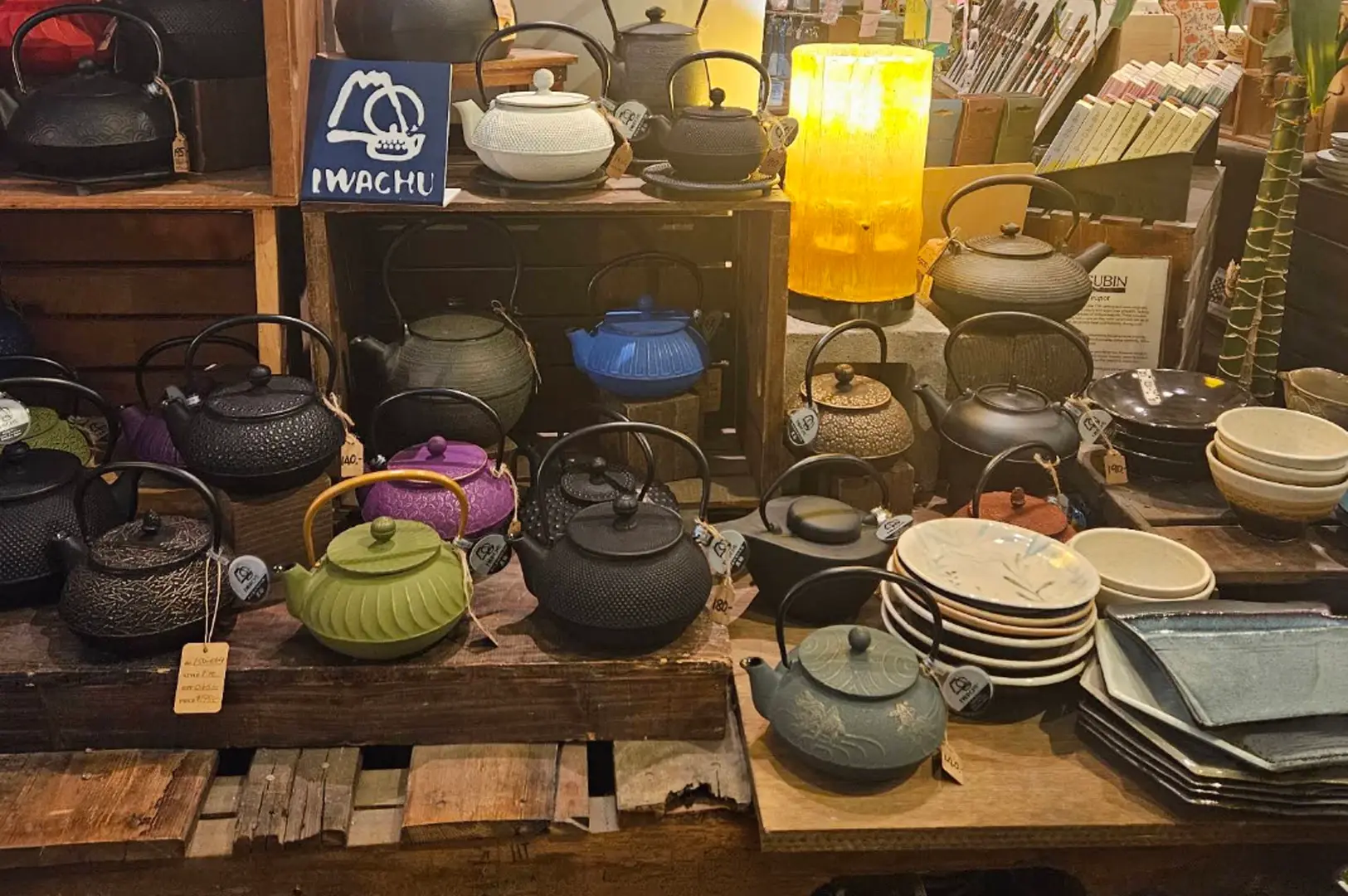 A wide shot of a retail display shelf filled with Japanese-style cast iron teapots, known as tetsubin, in various colors including black, blue, green, and purple. The shelves are rustic wood, and some items feature original packaging or labels. A bright, yellow glowing lamp sits on the middle shelf, illuminating nearby ceramics.