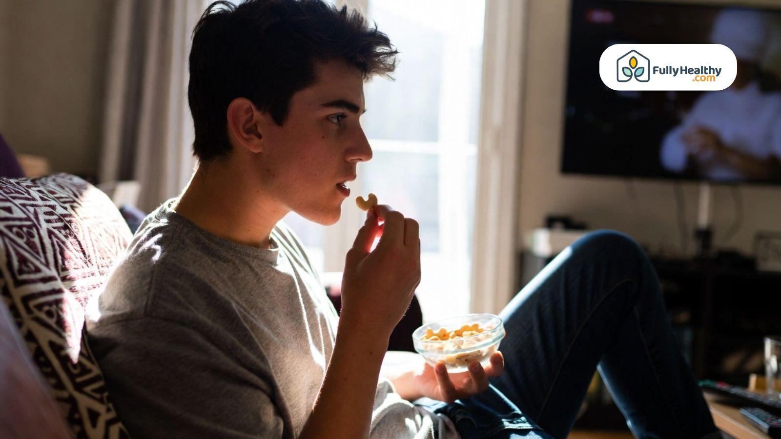 Person sitting on a couch eating nuts from a small bowl in a relaxed home setting.