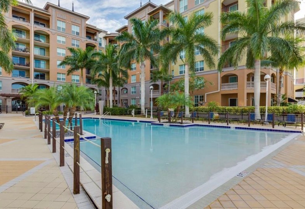 The image features a luxurious riverfront condominium building with modern architecture, situated along the scenic Caloosahatchee River in downtown Fort Myers. Towering palm trees surround the property, which includes amenities like a resort-style pool and fitness center, making it an attractive option for prospective properties consumers seeking real estate in the Alta Mar community.
