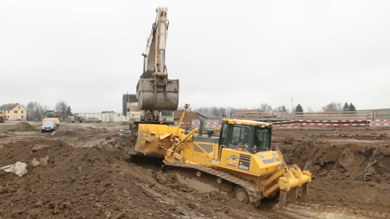 A large yellow bulldozer and excavator working on a construction site, moving dirt and digging a trench with houses and barriers in the background