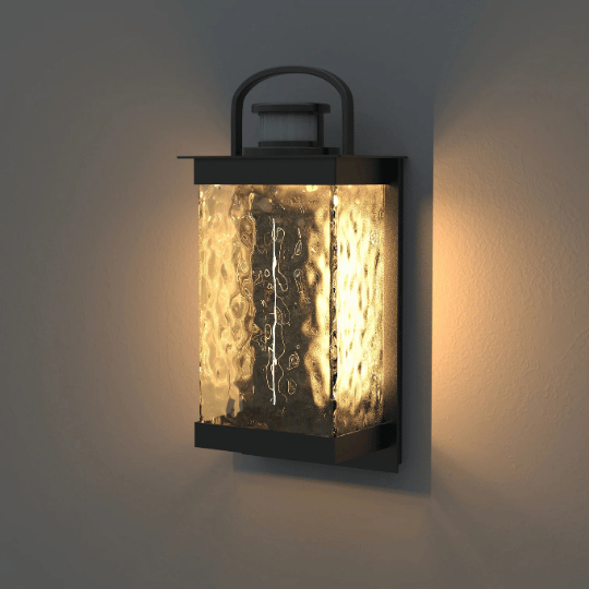 Wall-mounted outdoor lantern with black frame and textured glass, emitting a warm ambient glow against a light-colored wall.