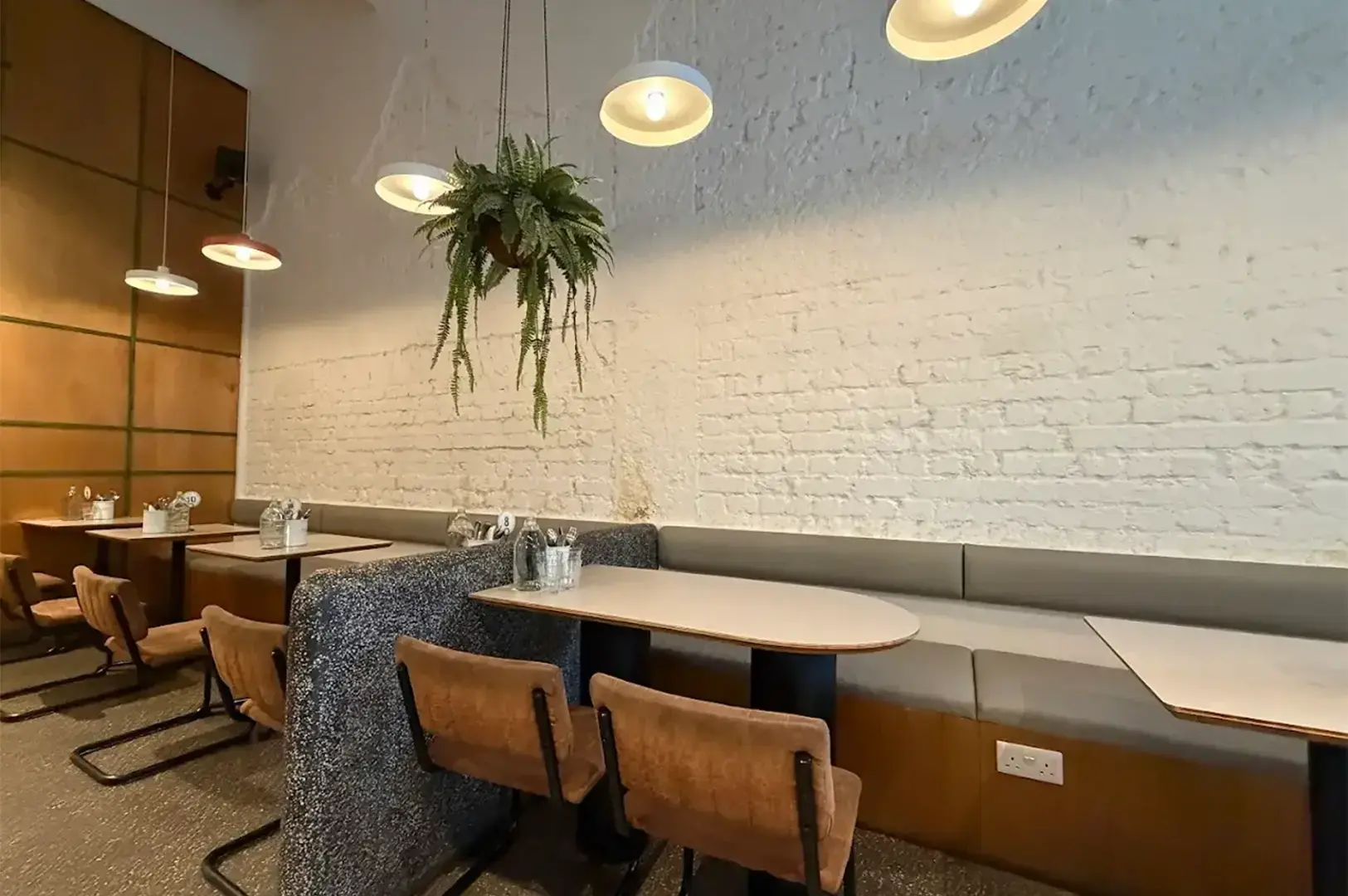 Chic café interior with wooden tables, brown chairs, hanging ferns, and warm pendant lights. Textured white brick wall adds a cozy atmosphere.