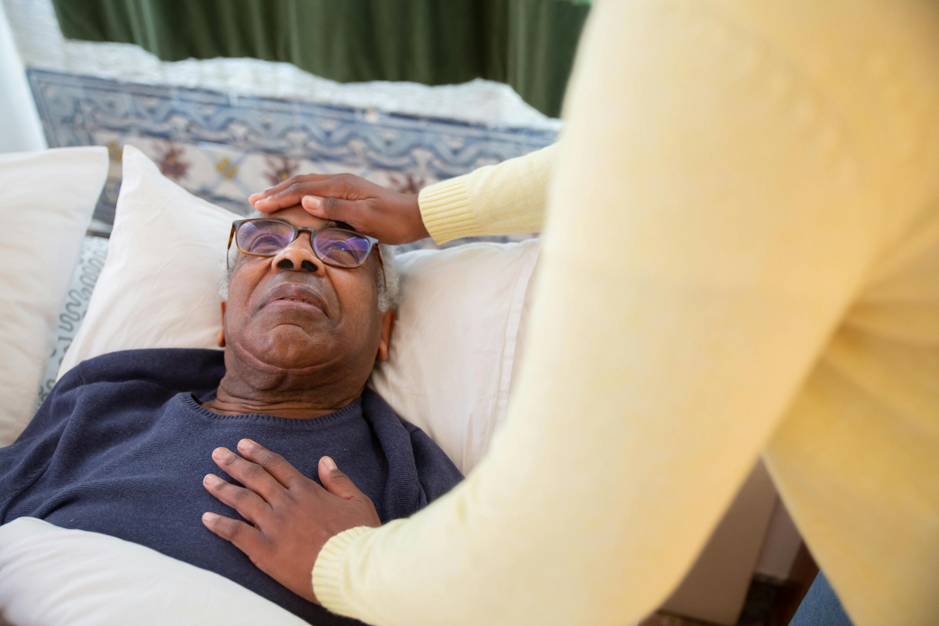 An elderly man in bed with someone's hands on his forehead and chest to comfort him.