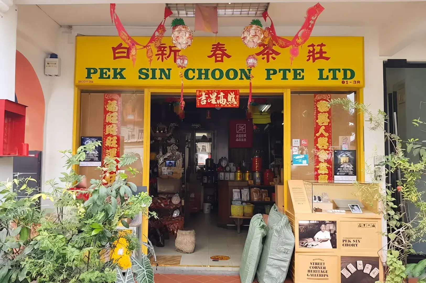 Storefront of "PEK SIN CHOON PTE LTD" tea shop with a yellow sign, Chinese New Year decorations, and potted plants.
