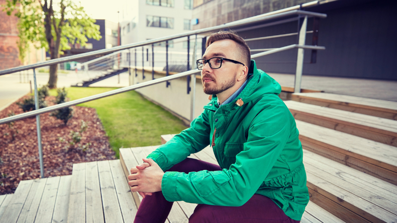 A person in glasses and a green Gorpcore jacket sits on wooden steps outside a building.
