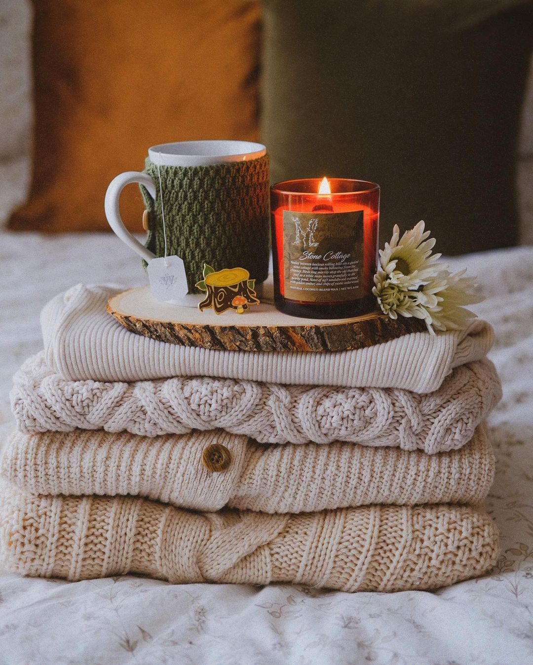 A pile of cozy knit sweaters in creamy off whites folded neatly on a bed with a candle and tea cup on top.