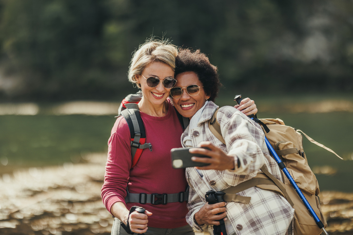 Two mature women in sunglasses stopping to snap a selfie while hiking. 