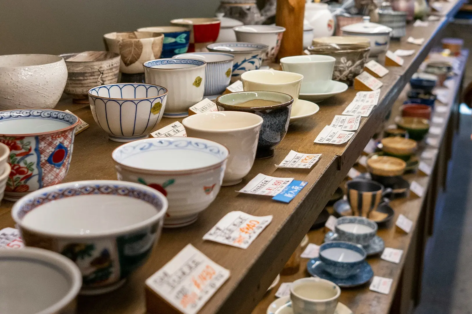 A shallow depth-of-field view of shelves in a teaware shop, stocked with a variety of ceramic bowls and teacups. The items are arranged in rows, with many featuring hand-written price tags placed in front of them. The foreground shows bowls with intricate blue patterns and plain white finishes, while the background reveals an array of textured, patterned, and solid-colored vessels.