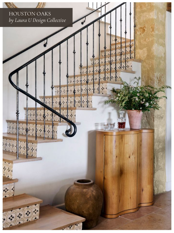 A rustic entryway featuring a staircase with decorative tile risers and black iron railings. A curved wood cabinet and large ceramic pot sit beneath the stairs, accented with greenery.