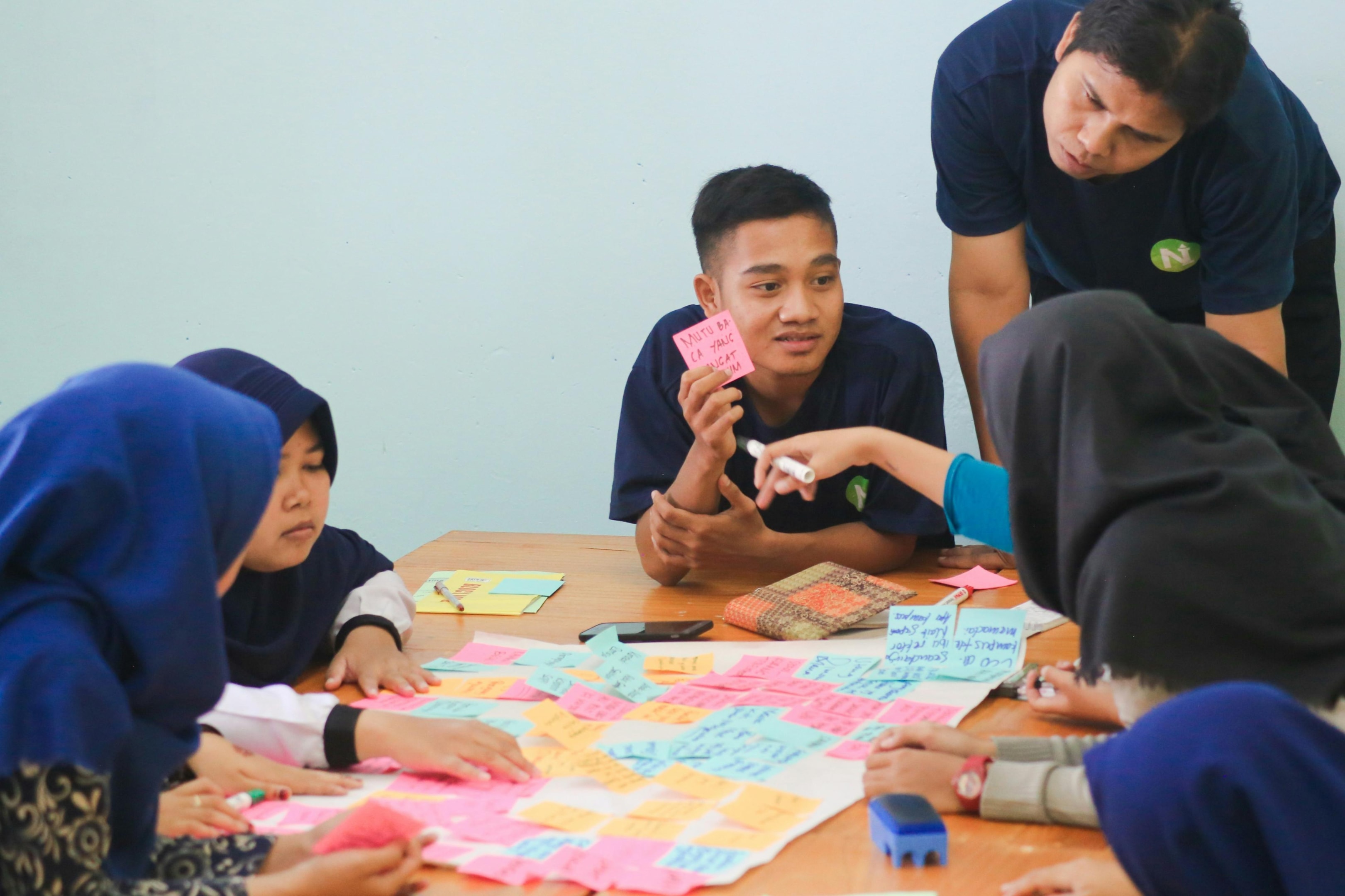 This image captures a lively collaborative brainstorming session where a group of young adults gathers around a table covered in colorful sticky notes. A standing facilitator observes the interaction as one participant holds up a note for discussion, highlighting an environment of active idea sharing and teamwork.