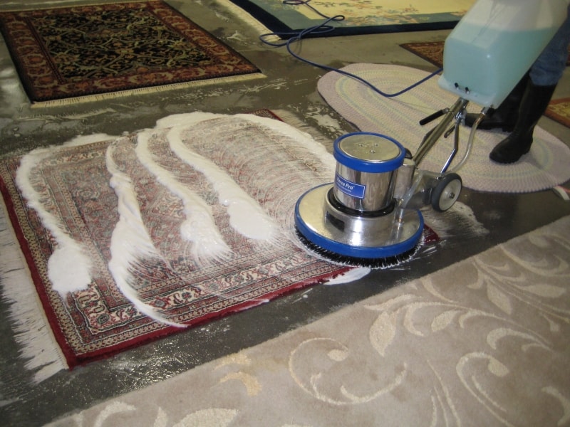 A professional rug cleaning machine with a blue rotary brush scrubbing a small oriental rug covered in cleaning foam. The setup is in a workshop environment with multiple other rugs visible in the background on a concrete floor.