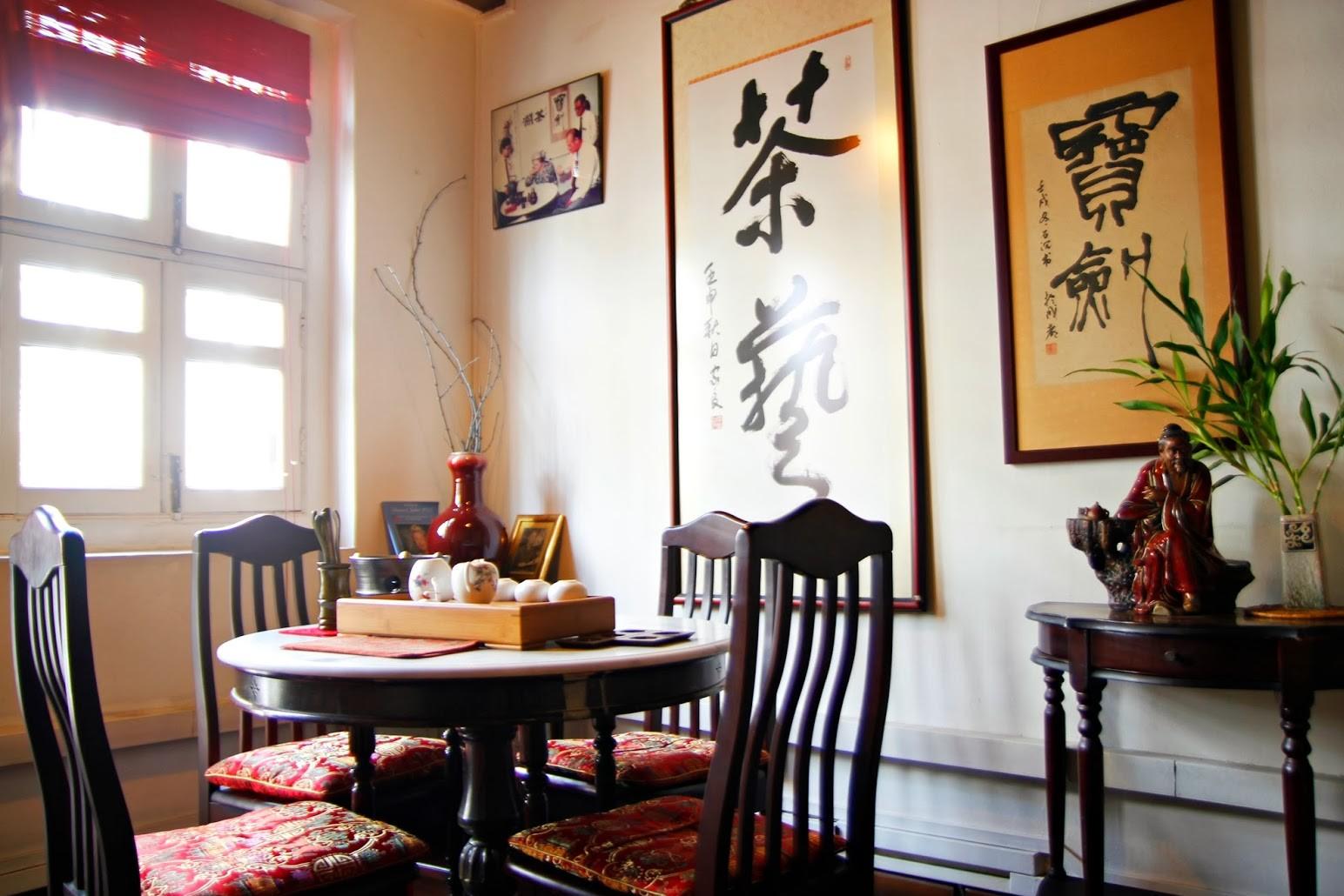 This traditional tea room features a round wooden table set with a tea service and chairs with vibrant red cushions. Large vertical scrolls with Chinese calligraphy and a decorative figurine accent the warm, sunlit space.