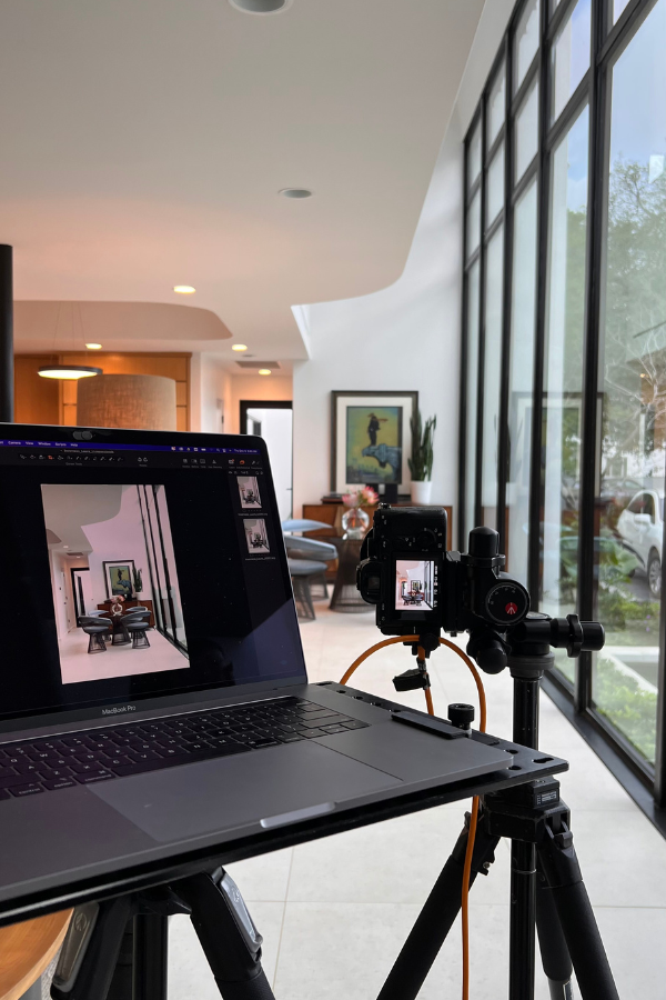 A photography setup shows a laptop displaying interior images connected to a camera on a tripod, facing a bright bathroom space in the background.
