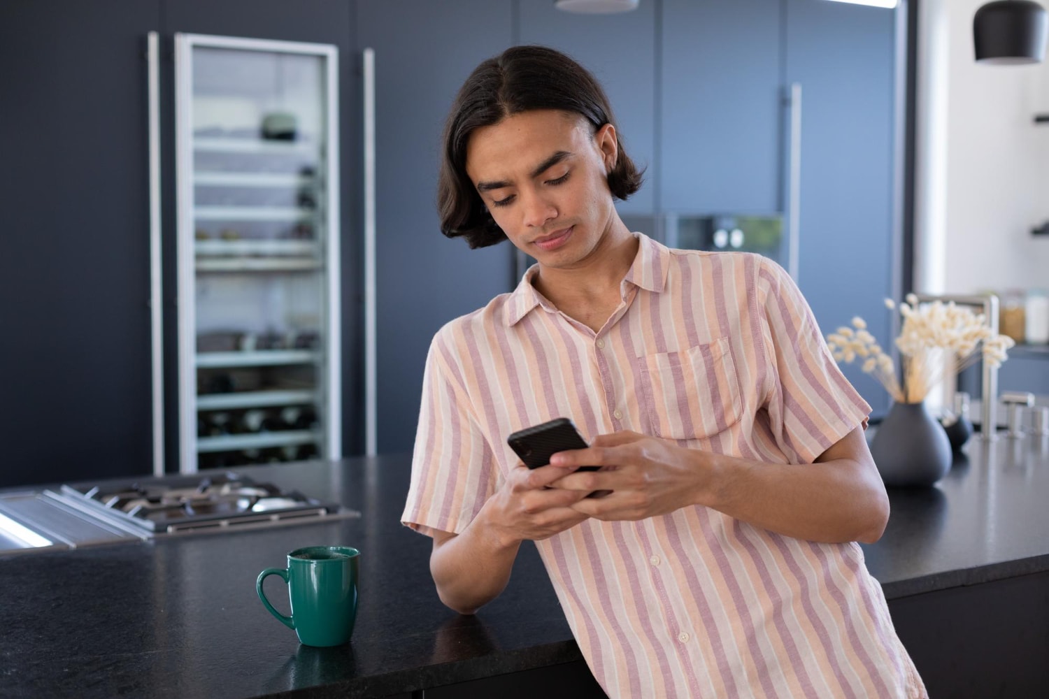 A young man leans against a kitchen counter with a coffee mug nearby, reading something on his smartphone. This casual indoor scene emphasizes how internal business messaging can seamlessly integrate into daily workflows via business SMS.