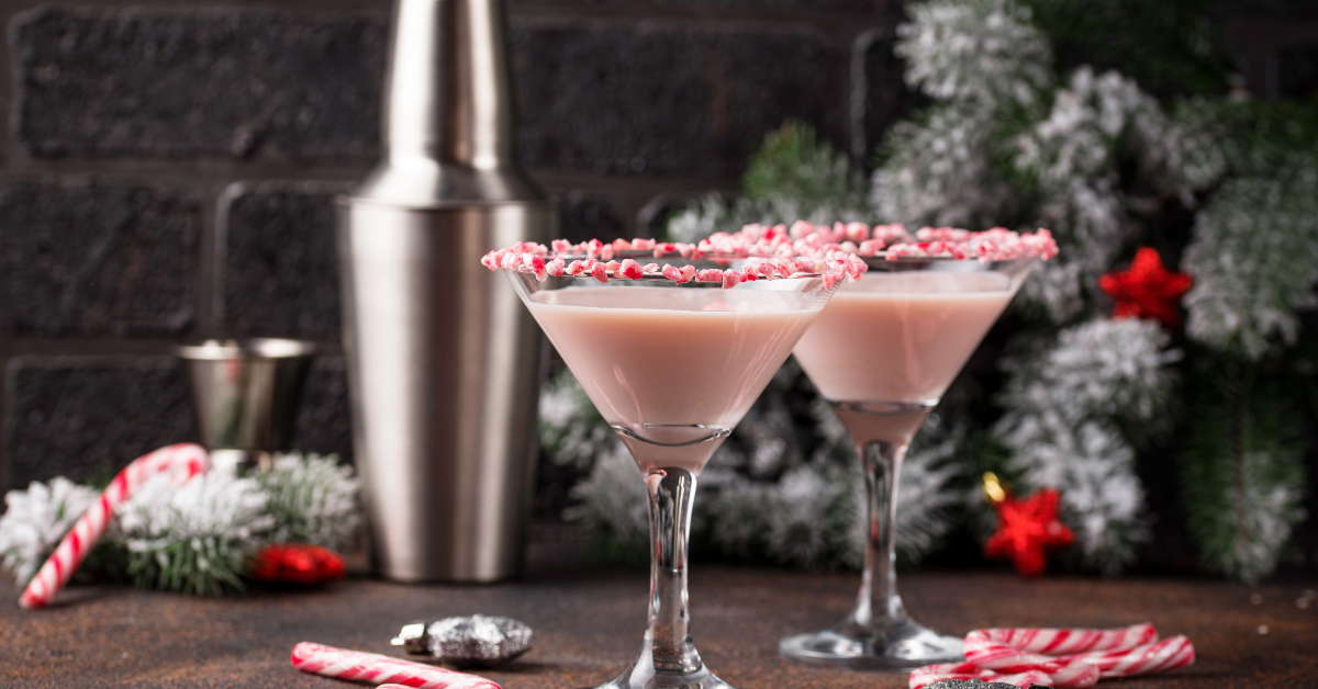 Two pink peppermint martinis with crushed candy cane rims sit in front of a stainless steel cocktail shaker, surrounded by candy canes, holiday decorations, and frosted greenery, creating a festive winter cocktail scene.