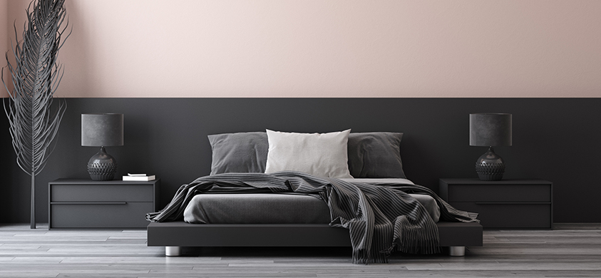 This minimalist bedroom setting features deep, dark grey furnishings that are mainly rectangular with straight, clean lines. There is little decoration beyond two textured bedside lamps, a dark grey throw blanket, and a feather-like sculpture in the left corner. The room has a mostly greyscale palette, besides a pale grey/pink colour covering the upper half of the back wall.