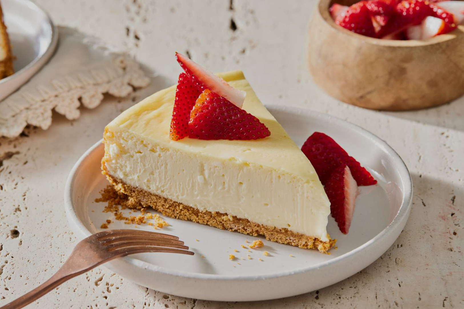 A creamy slice of cheesecake with a golden crust is garnished with fresh sliced strawberries on a white plate, next to a cup of additional strawberries.