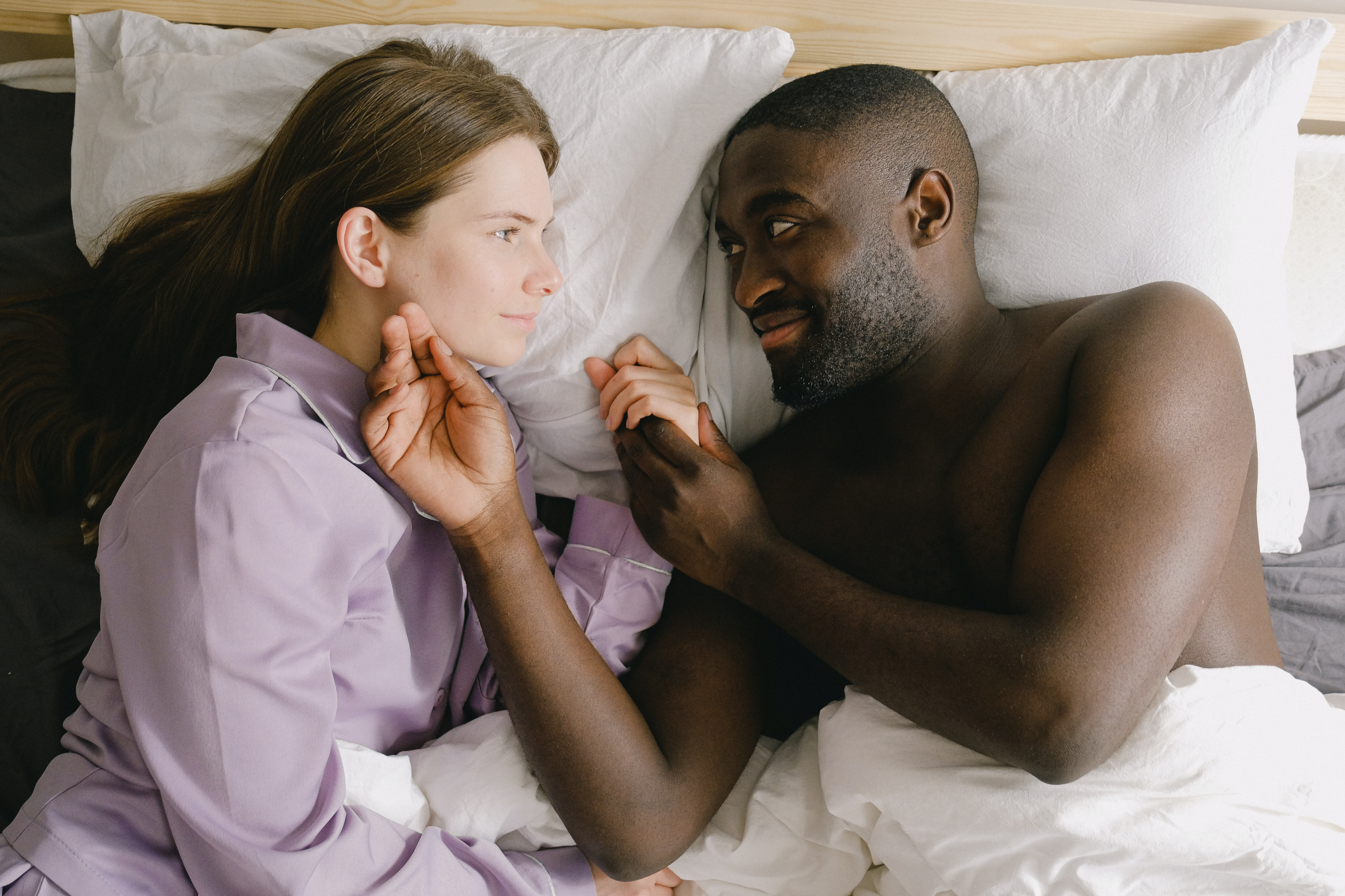 Couple lay in bed, facing each other, holding hands.