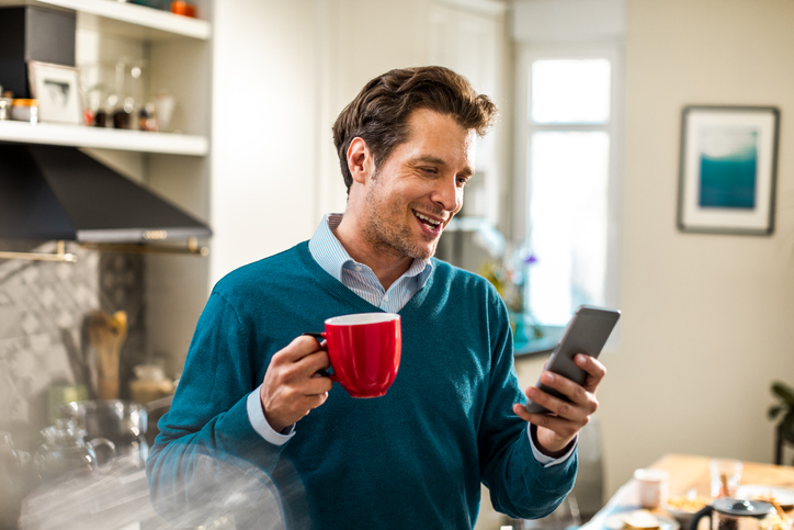 Dark haired man in a teal sweater holding a red coffee cup with a cell phone.