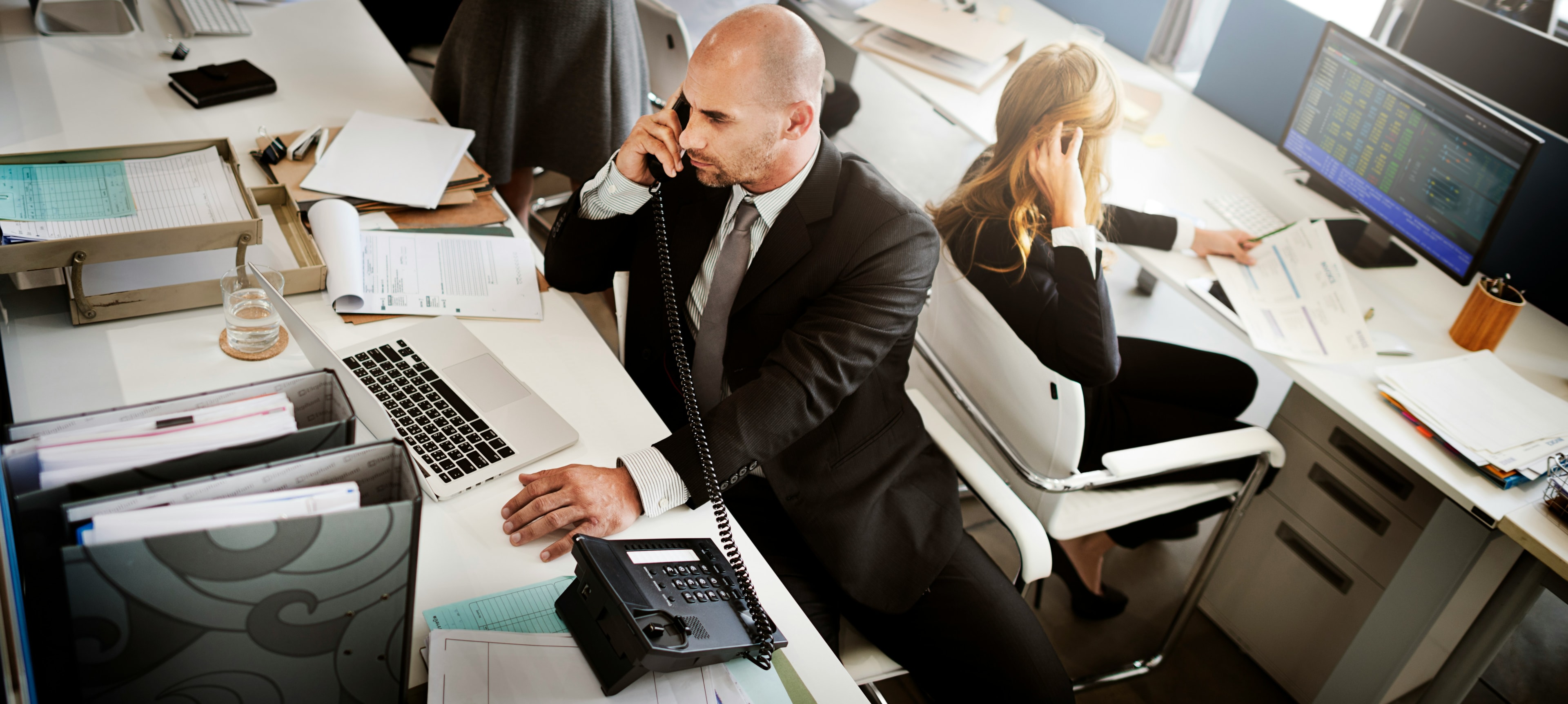 Bald man taking a call in an office, woman at desk behind him