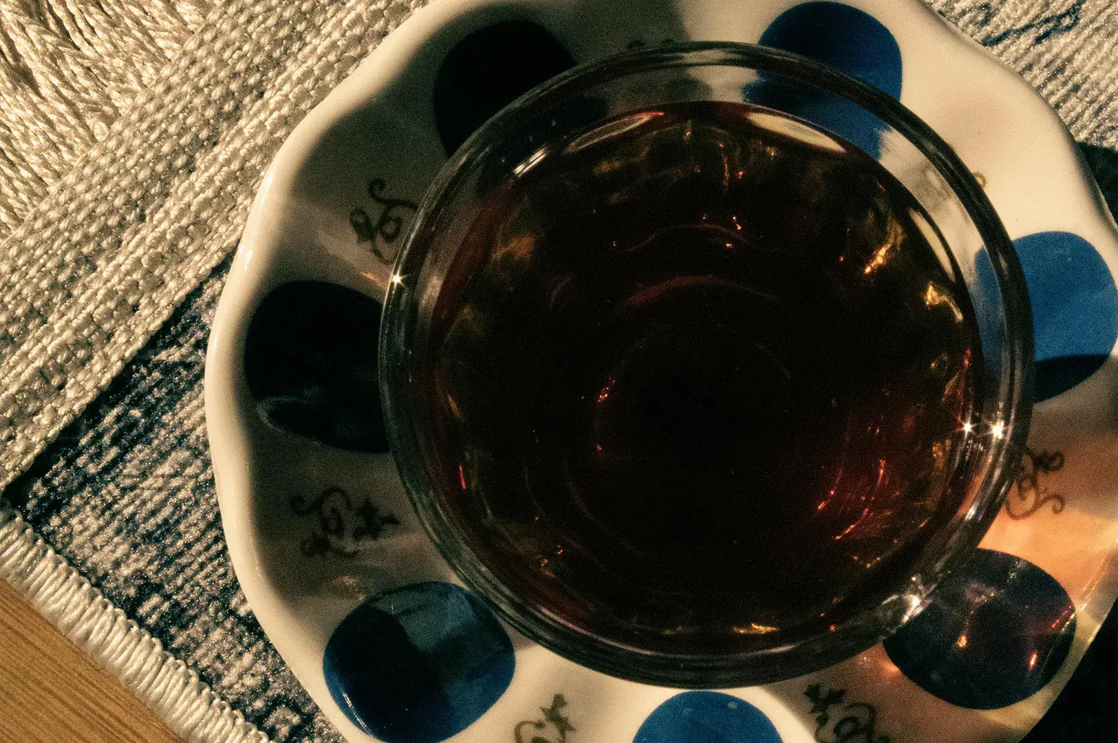 A high-angle, close-up view features a dark-colored beverage in a small, clear glass cup. The cup rests on a decorative saucer adorned with a repeating pattern of blue circles and delicate, dark line art, all placed upon a textured, patterned fabric mat.