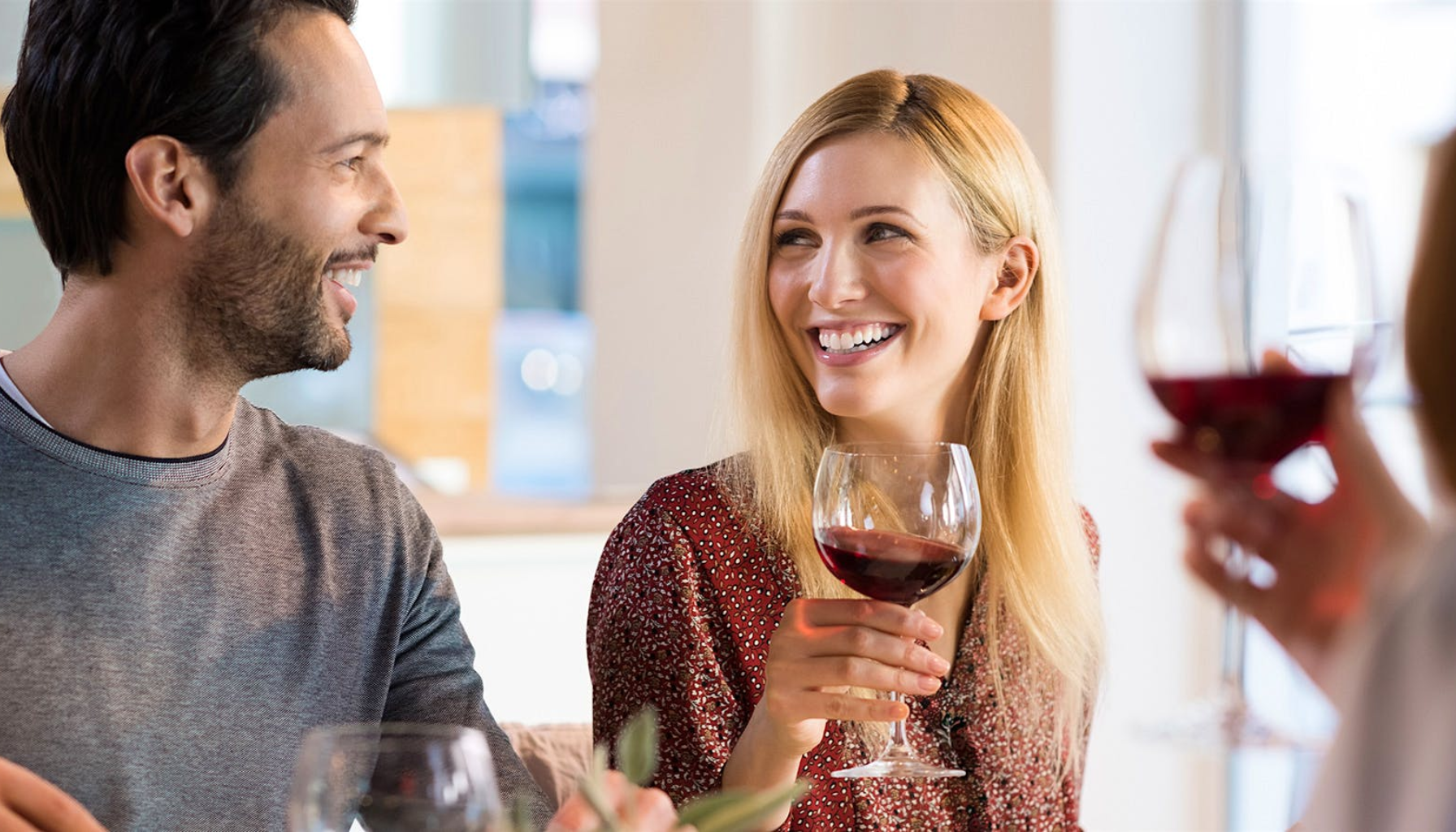 To personer smiler og nyter et glass rødvin i et lyst miljø.